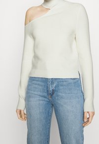 Off-white ribbed sweater with one shoulder exposed, high neckline, and fitted long sleeves. Paired with blue denim jeans.