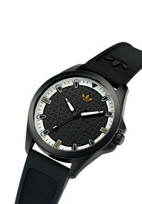 Montre de sport noire avec un cadran texturé, des marqueurs d'heure blancs et un bracelet en silicone noir ; présente un accent de logo doré et des détails argentés.