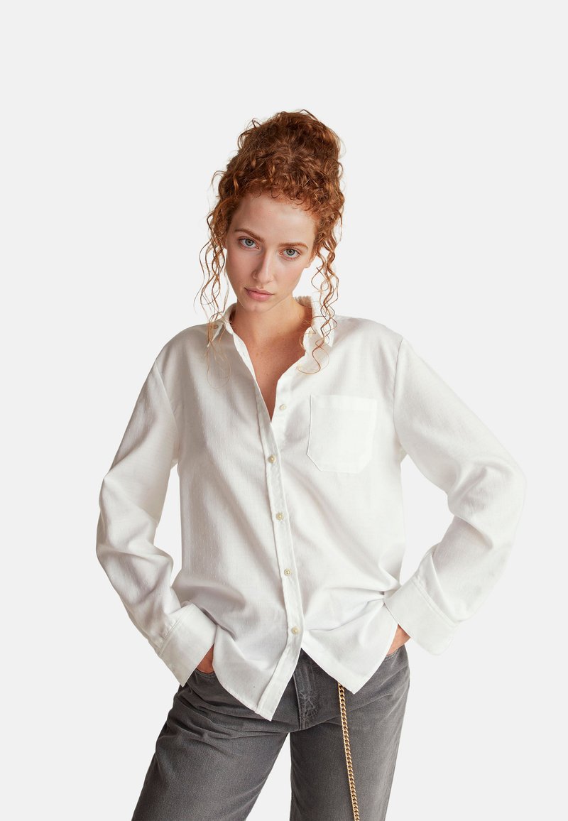 Camicia bianca con bottoni, tasca sul petto, tessuto strutturato e maniche lunghe. Abbinata a pantaloni grigi con dettaglio a catena.