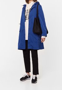 Blå vattentät parka med huva, tryckknappar och sidofickor över en ljus hoodie, kombinerat med svarta cropped jeans och svarta sneakers.