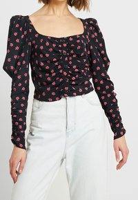 Top lungo a maniche nere con fiori, spalle a sbuffo, dettagli arricciati e chiusura con bottoni. Presenta piccoli motivi di rose rosa su fondo scuro.