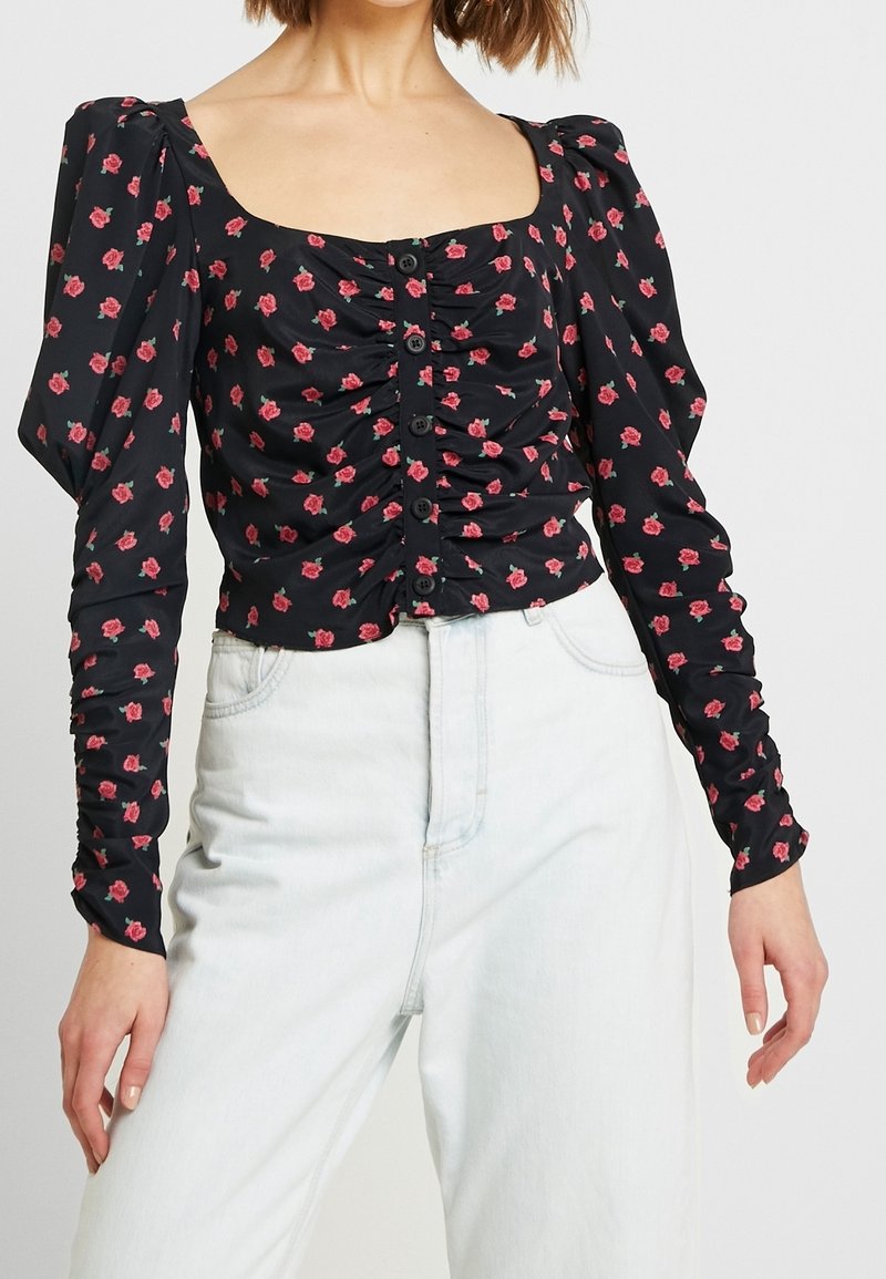 Top lungo a maniche nere con fiori, spalle a sbuffo, dettagli arricciati e chiusura con bottoni. Presenta piccoli motivi di rose rosa su fondo scuro.