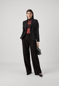 Zwarte op maat gemaakte blazer over een doorschijnende, gestreepte rode en zwarte top, gecombineerd met wijde zwarte broek en een kleine zwarte handtas. Fijne texturen.