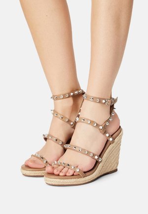 Sandales compensées beiges avec des brides cloutées et un anneau de orteil, dotées d'une semelle en jute tissée. Texture lisse avec des accents métalliques pour les détails.