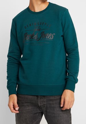 Mężczyzna ubrany w morski sweter Jack & Jones z czarnym napisem i ciemnoszare jeansy, stojący na jednolitym jasnym tle.
