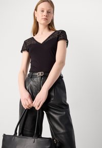 Top noir ajusté avec des manches en dentelle, décolleté en V. Associé à un pantalon en cuir noir taille haute et un sac fourre-tout noir.