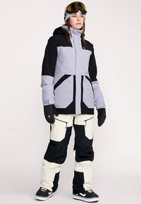 Ski jacket in light purple and black with geometric color blocking; paired with cream and black pants, gloves, and ski goggles.