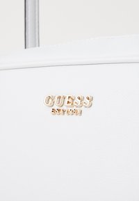 Bolso de mano texturizado en blanco con un logo dorado de "GUESS" y un detalle de "EST 1981" en la parte frontal. Material sintético suave con un diseño elegante.