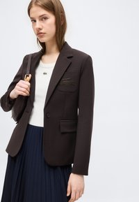 Jeune femme portant un blazer sombre avec un emblème de crocodile, un haut blanc, une jupe plissée bleu marine, et portant un sac à bandoulière sombre sur fond gris.