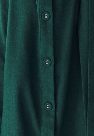 Donkergroene shirt met stoftextuur, voorzien van vijf ronde knopen in zilver. De compositie benadrukt de knopensluiting en plooien.