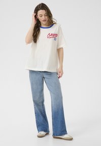 Cream-colored t-shirt with blue neckline and red collegiate print. Paired with light blue wide-leg jeans. Shoes are beige and white.