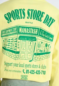 Camiseta de manga corta en amarillo brillante con texto gráfico verde e ilustraciones de equipo deportivo, promoviendo un evento de una tienda de deportes local.
