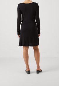 Czarna sukienka z długim rękawem o dopasowanej sylwetce, okrągłym dekolcie i detalem wiązania z tyłu. Gładka tekstura i długość do połowy łydki, zestawiona z czarnymi słupkowymi szpilkami.