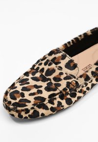 Chaussure à enfiler à motif léopard avec une surface douce et texturée, dotée d'un col matelassé et d'une semelle robuste. La palette de couleurs comprend du beige, du noir et du marron.