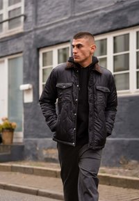 Quiltet sort jakke med en brun krave og to brystlommer, båret over en sort hættetrøje og grå sweatpants, gående på en brostensbelagt gade.