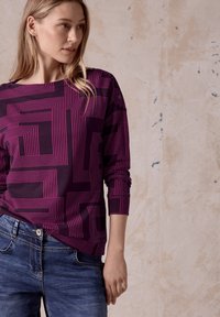 Top de manga larga púrpura con líneas geométricas rosas, escote redondo y puños acanalados, combinado con jeans de mezclilla azules, presentado sobre un fondo neutro.