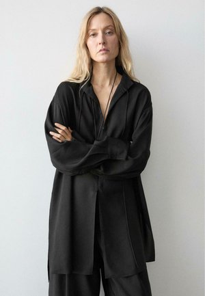 Kvinde med langt blondt hår iført løs sort skjorte og bukser står med armene krydset mod en ensartet lys baggrund.