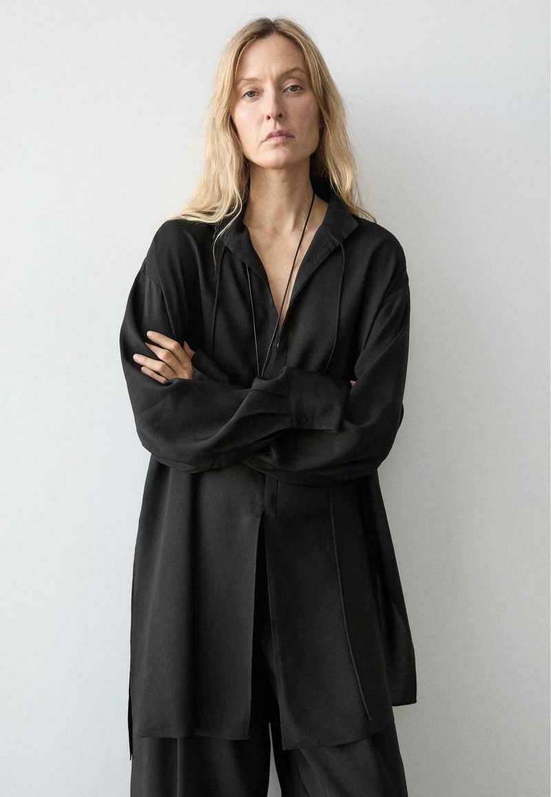 Frau mit langen blonden Haaren, die ein lockeres schwarzes Hemd und Hose trägt, steht mit verschränkten Armen vor einem schlichten hellen Hintergrund.