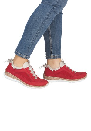 Person trägt eng anliegende blaue Jeans und rote Turnschuhe mit weißen Schnürsenkeln und weißen Sohlen und steht auf weißem Hintergrund.