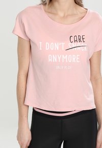 Žena v růžovém tričku s nápisem "Už mi to je jedno," kde slovo "je jedno" je napsáno přes přeškrtnuté slovo "svačina," a černé legíny.