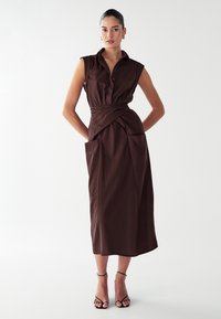 Robe marron sans manches avec un élégant devant boutonné, une ceinture à la taille et des poches latérales. L'ourlet tombe sous le genou. Portée avec des talons noirs à lanières.