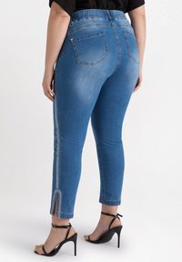 Vaqueros azules de corte slim, con un detalle de cremallera en el lateral y una abertura en el tobillo, combinados con sandalias negras de tacón alto.