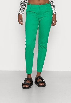 Partie inférieure du corps d'une personne portant un pantalon slim vert vif avec une ceinture marron et des sandales noires à double sangle, sur un fond blanc uni.