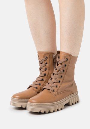 Bottines en cuir marron clair à lacets avec fermetures éclair latérales et semelles en caoutchouc beige épaisses portées sur jambes nues sur un fond clair uni.