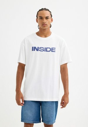 Camiseta blanca de algodón con un audaz texto en navy "INSIDE" en el pecho. Corte relajado con mangas cortas, combinada con pantalones cortos de mezclilla.