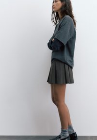 Młoda kobieta stojąca bokiem, ubrana w szary warstwowy top, plisowaną spódnicę, pasiastych skarpetkach i czarne loafersy na jednolitym tle.