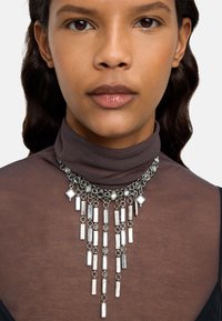 Srebrny naszyjnik składający się z wielu kaskadowych prostokątnych i diamentowych elementów metalowych; zawiera teksturowane i gładkie powierzchnie. Noszony na brązowej bluzce.