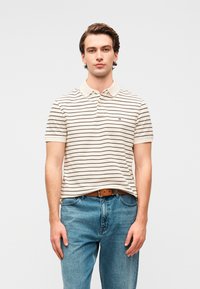 Πόλο μπλούζα κατασκευασμένη από βαμβάκι, με οριζόντιες ρίγες σε μπεζ και καφέ χρώμα, γιακά και μικρό λογότυπο στο στήθος. Φοριέται με μπλε τζιν.