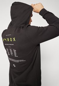 Melns kapucis ar auklu. Aizmugurē ir baltas krāsas teksti un grafikas, tostarp "HYROX" neona dzeltenā krāsā. Mīksta auduma, brīvs piegriezums.