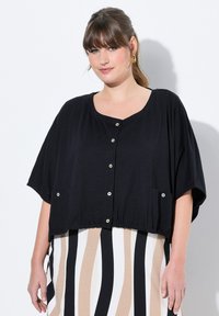 Cropped top nero con maniche corte, caratterizzato da un scollo a girocollo, chiusura a bottone e orlo arricciato; indossato sopra una gonna a righe.