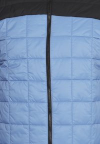 Giacca trapuntata con pannelli orizzontali blu chiaro e neri, caratterizzata da una zip nera che scorre verticalmente al centro.