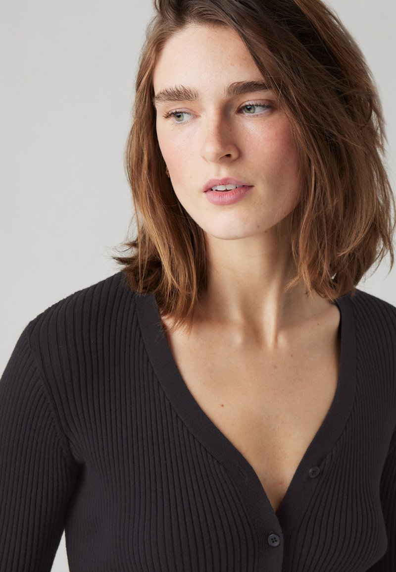 Schwarze, gerippte Strickjacke mit V-Ausschnitt, ausgestattet mit zwei Knöpfen und einer weichen Textur. Der Stoff wirkt leicht und figurbetont.