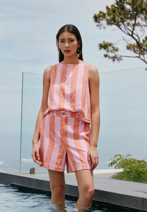 Femeie stând în apă lângă piscină, purtând un top fără mâneci cu dungi verticale roz și portocalii și pantaloni scurți asortați, cu cercei și copaci în fundal.