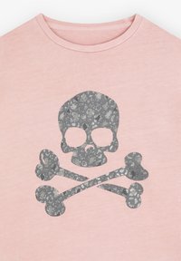 Camiseta de color rosa claro con un gráfico de calavera y huesos cruzados plateado brillante, que presenta un patrón texturizado dentro del diseño. Textura de tela suave.