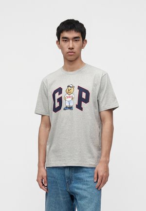 Giovane uomo che indossa una T-shirt grigio chiaro con grandi lettere blu navy "G" e "P" e un orso che indossa un outfit Gap, abbinato a jeans blu.