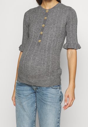 Femme portant un pull tricoté gris côtelé avec cinq grands boutons sur le devant et des manches courtes à volants, assorti à un jean bleu clair.