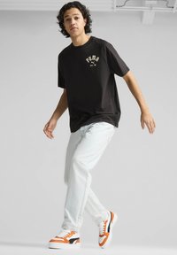 Sort t-shirt med korte ærmer og guld "PUMA" logo, lyseblå jeans og hvide sneakers med orange og sorte detaljer.