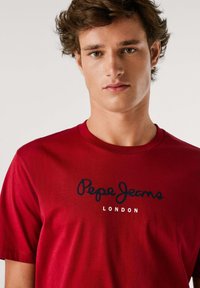 Raudona medvilninė marškinė su apvalia iškirpte ir trumpomis rankovėmis, su juodu „Pepe Jeans London“ logotipu atspausto per krūtinę.