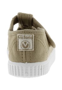 Zapato slip-on de lienzo color marrón claro con suela de goma blanca, que presenta una etiqueta rectangular con el logotipo en el talón. Tejido texturizado y diseño sencillo.