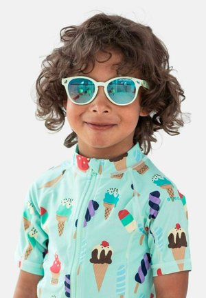 Smilende barn med krøllet hår iført lyseblå is-mønstret skjorte og reflekterende turkis solbriller.