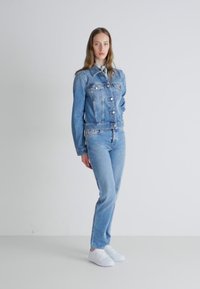 Light blue denim jacket with two chest pockets and metal buttons, paired with matching light blue jeans and white sneakers. Smooth texture.