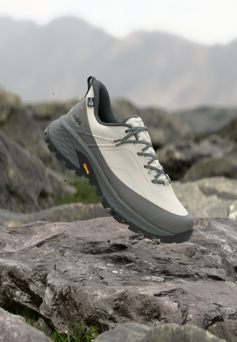 Ανοιχτό γκρι και σκούρο γκρι πεζοπορικό παπούτσι με σόλα Vibram, να αιωρείται πάνω από βραχώδες υπαίθριο έδαφος με θολά βουνά στο φόντο.