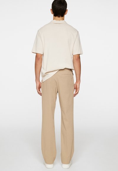 Beige Poloshirt mit kurzen Ärmeln, kombiniert mit beigen weit geschnittenen Hosen. Das Outfit zeichnet sich durch einen strukturierten Stoff und einen lässigen Schnitt aus.