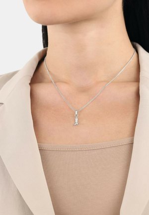 Zilveren halsketting met een klein hangertje in de vorm van een hond. De ketting is delicaat en rust boven een lichtbeige top, bedekt door een roomwitte blazer.