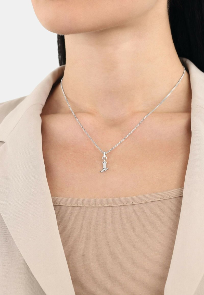 Collana d'argento con un piccolo pendente a forma di cane. La catena è delicata, poggiata su una maglia di un leggero beige, coperta da un blazer crema.