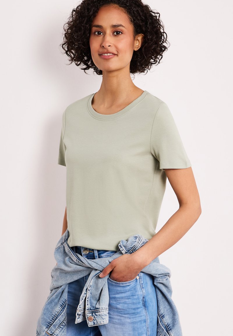 Femme aux cheveux bouclés portant un t-shirt vert clair à manches courtes et un jean bleu avec une veste en jean nouée autour de la taille.